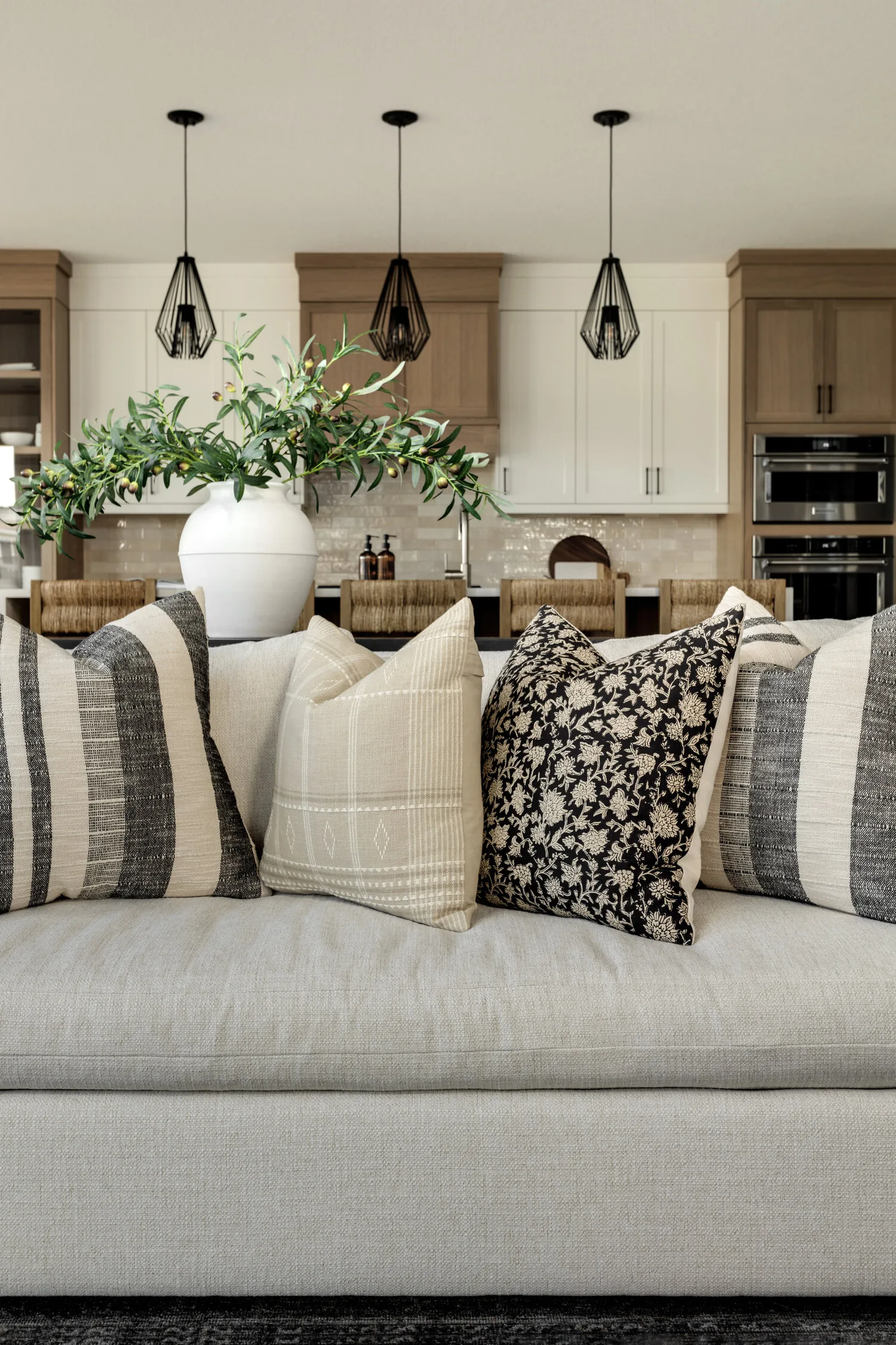 Layered sofa with patterned pillows, olive branches in a white vase, and a modern kitchen beyond